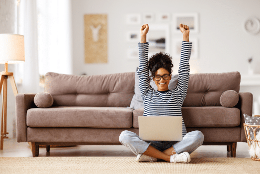Woman celebrating with arms raised, sitting on floor in front of couch with laptop