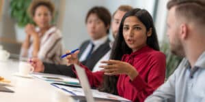 Photo of a meeting room with people sitting next to each other, and the focal point is of a South Asian woman speaking.